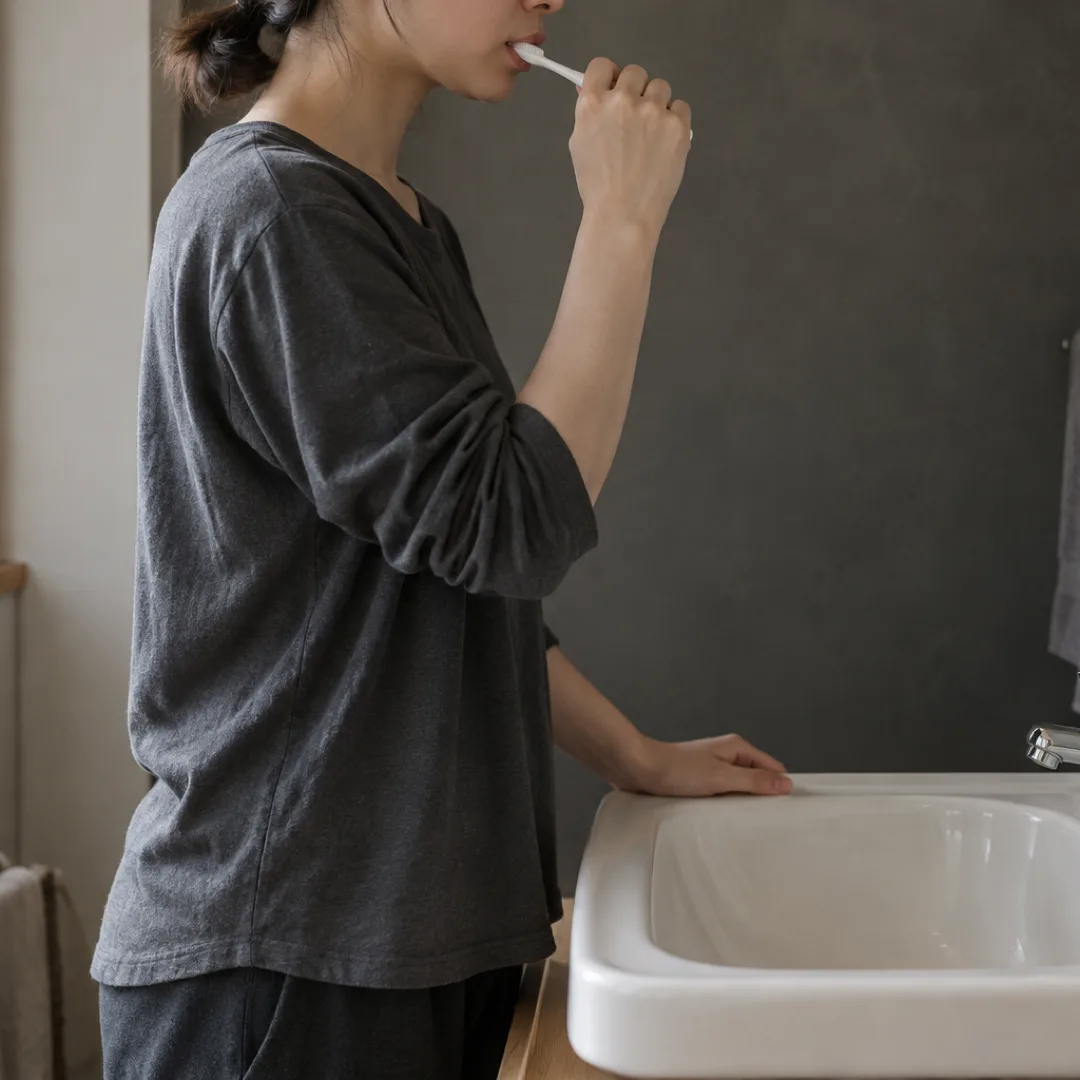 日本の住まいの洗面台で、チャコール色の服を着た大人が、口元にほとんど泡をつけずに歯を磨いている。