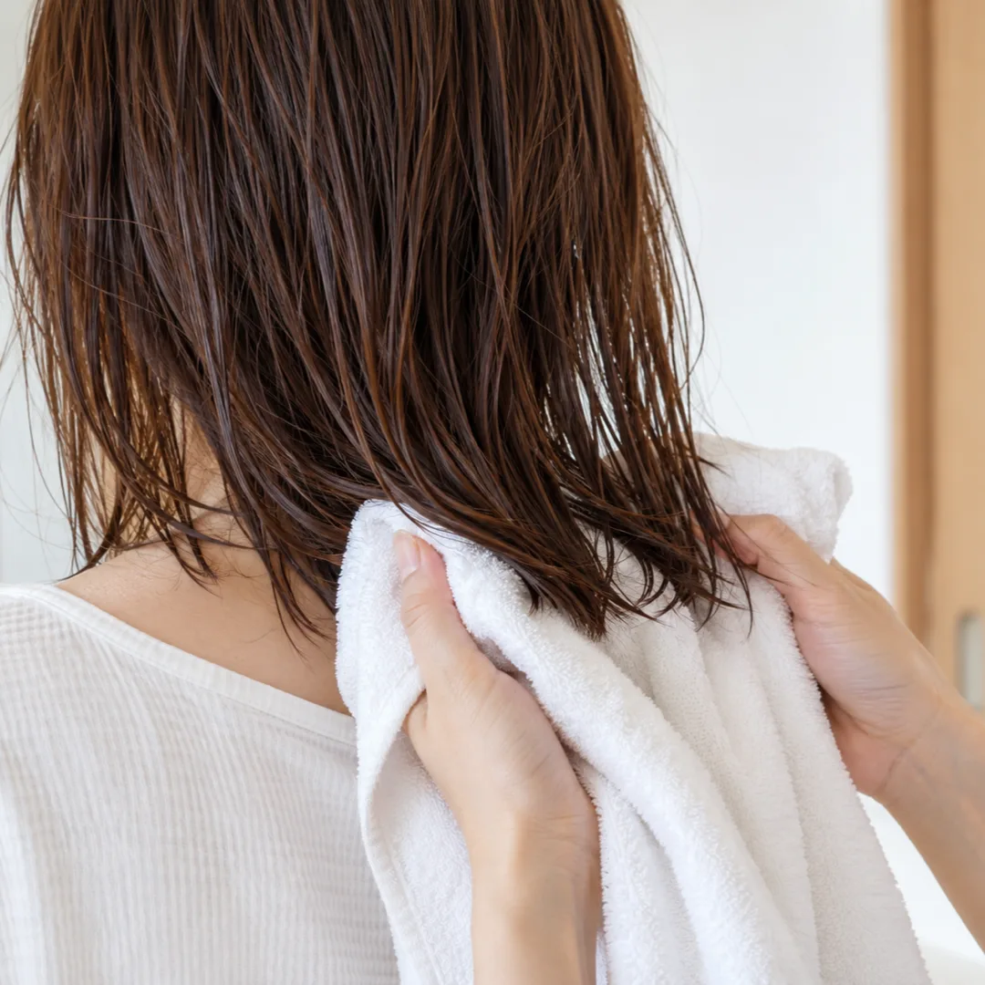 白い壁と淡い木の戸枠を背景に、濡れた栗色の髪の毛先を白いタオルでそっと押さえる人の後ろ姿。