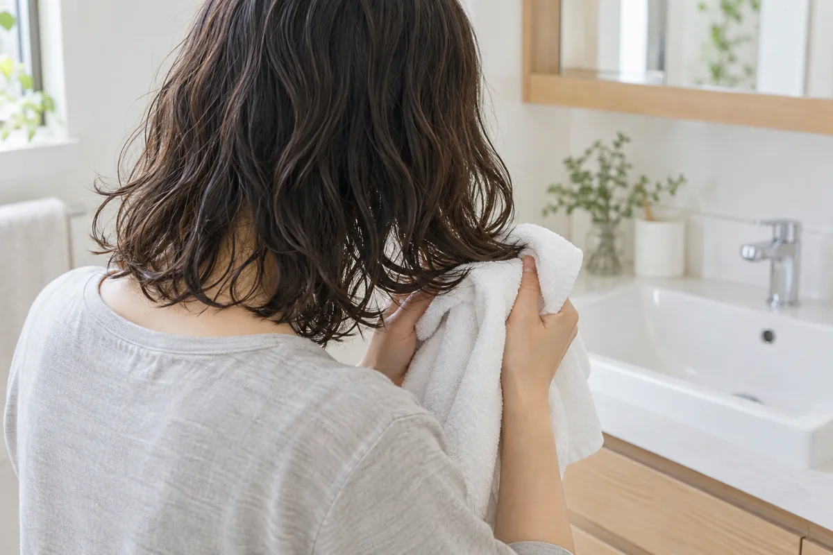 日本の洗面所で、ゆるいカールのある髪の毛先を白いタオルでそっと挟む人の後ろ姿。