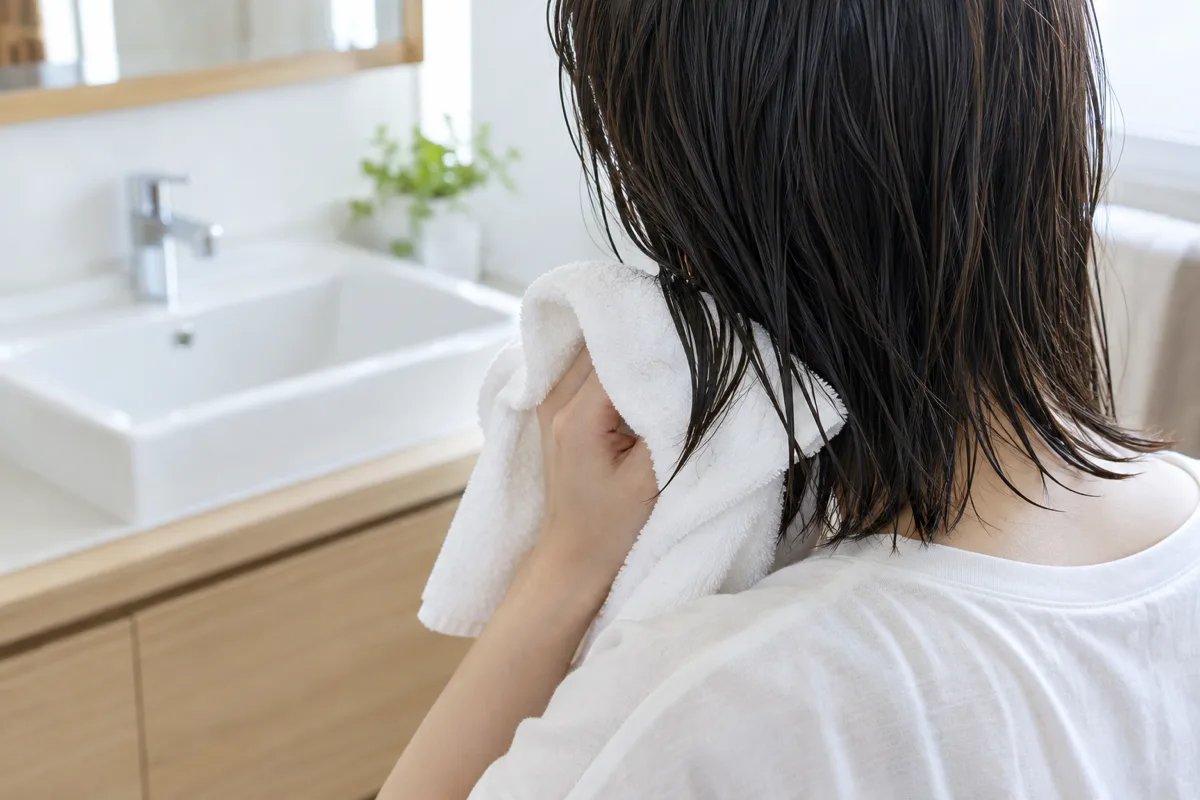 日本の洗面所で、白いタオルを使って濡れた黒髪の毛先を優しく押さえる人の後ろ姿。