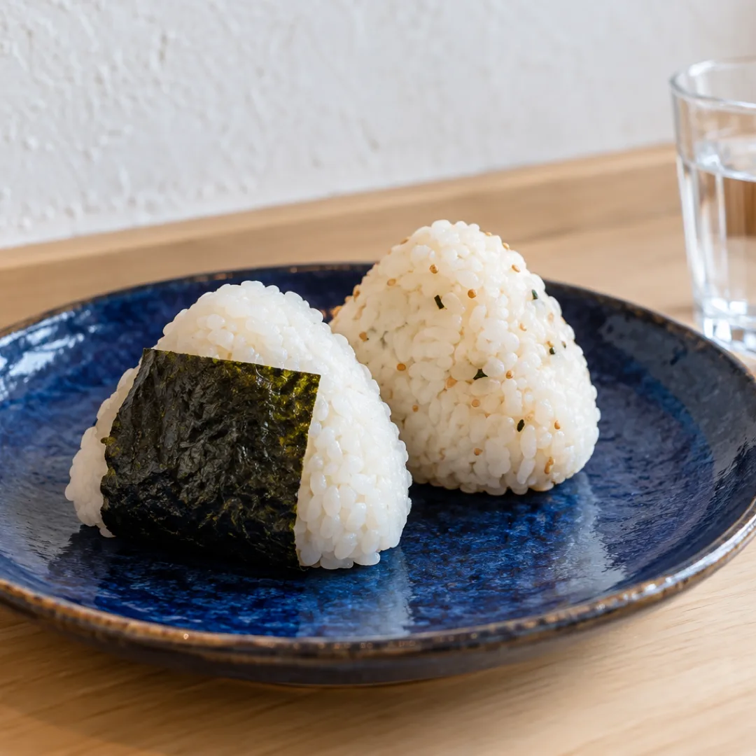 白い壁を背景に、淡い木のカウンターの藍色の皿におにぎりが二つ並び、隣に水の入ったグラスがあります。