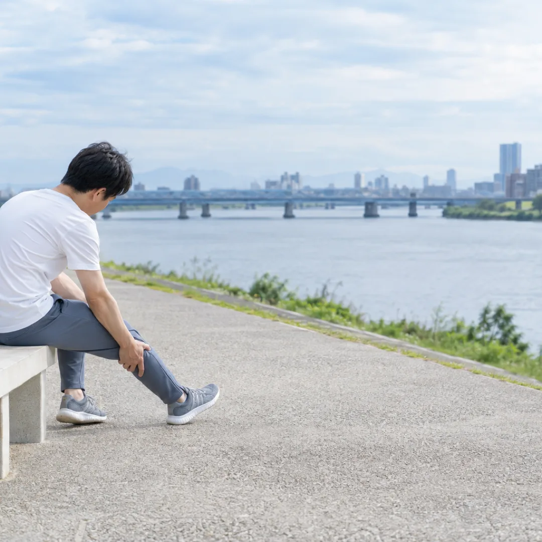 川沿いのベンチに座り、片脚を少し伸ばして両手でふくらはぎに触れる人。