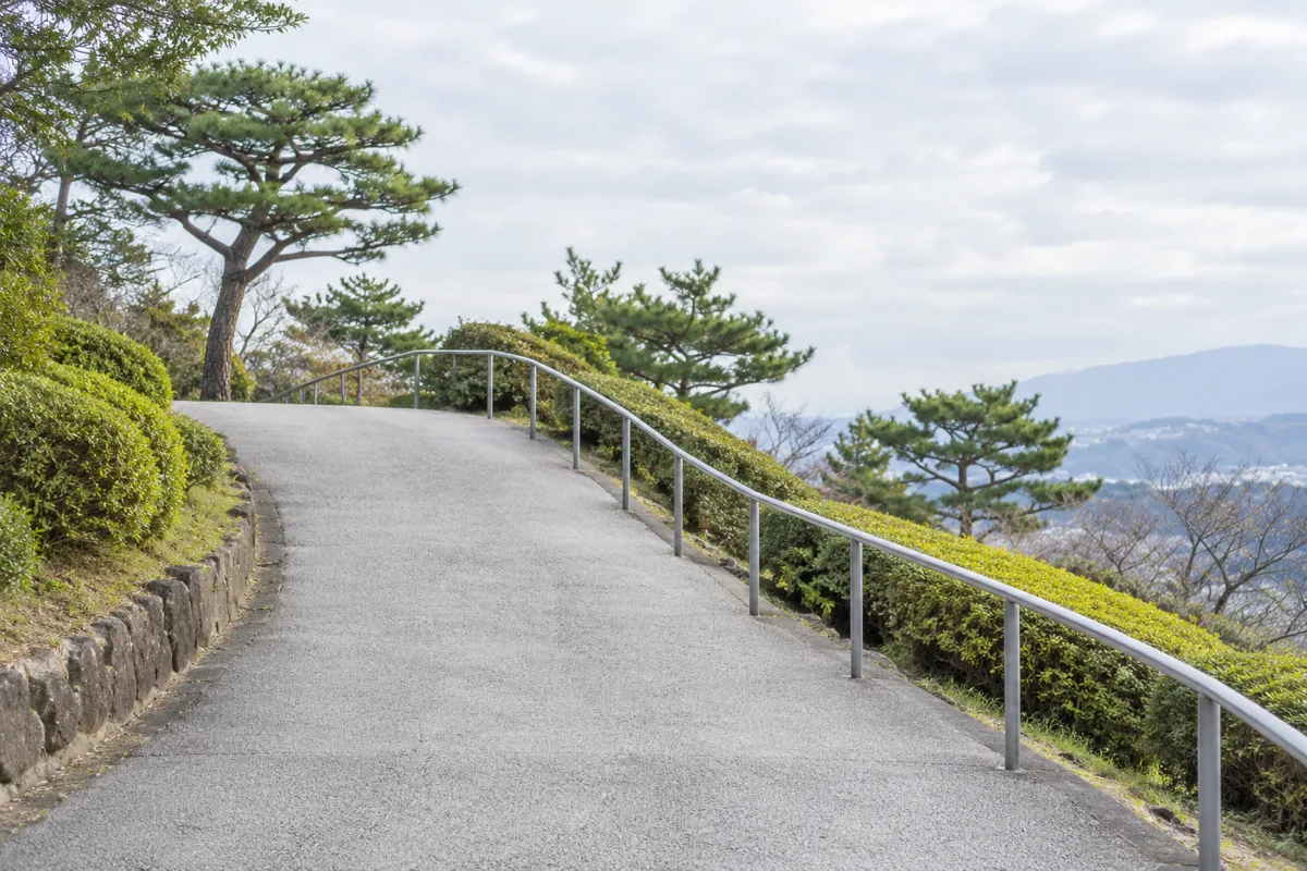 曇り空の下、日本の公園で、手すりに沿った灰色の歩道が緩やかに上り、頂上から下っている。
