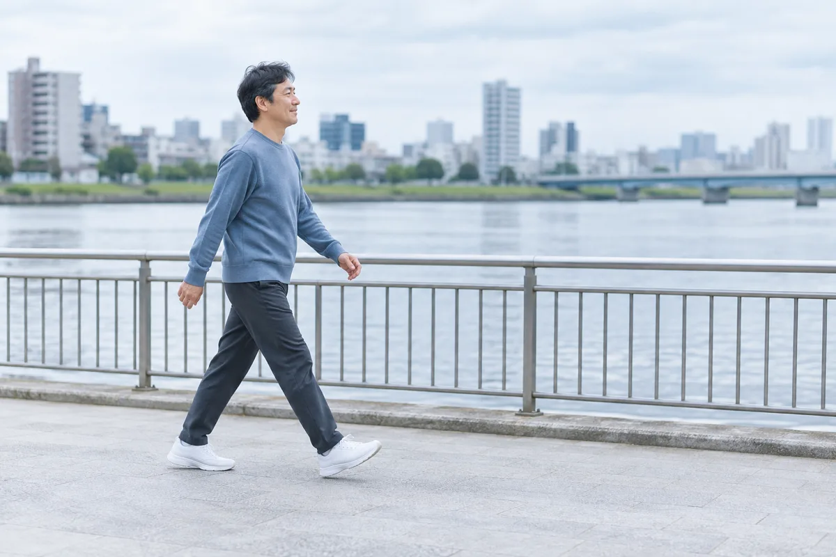 日本の川沿いの遊歩道を、青灰色の服と白い運動靴を身につけた大人が歩く横姿。