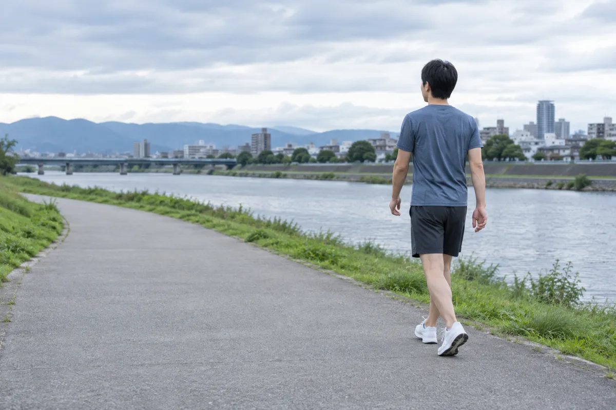 日本の川沿いの遊歩道で、青灰色の運動着を着た大人が腕を下ろしてゆっくり歩いている。