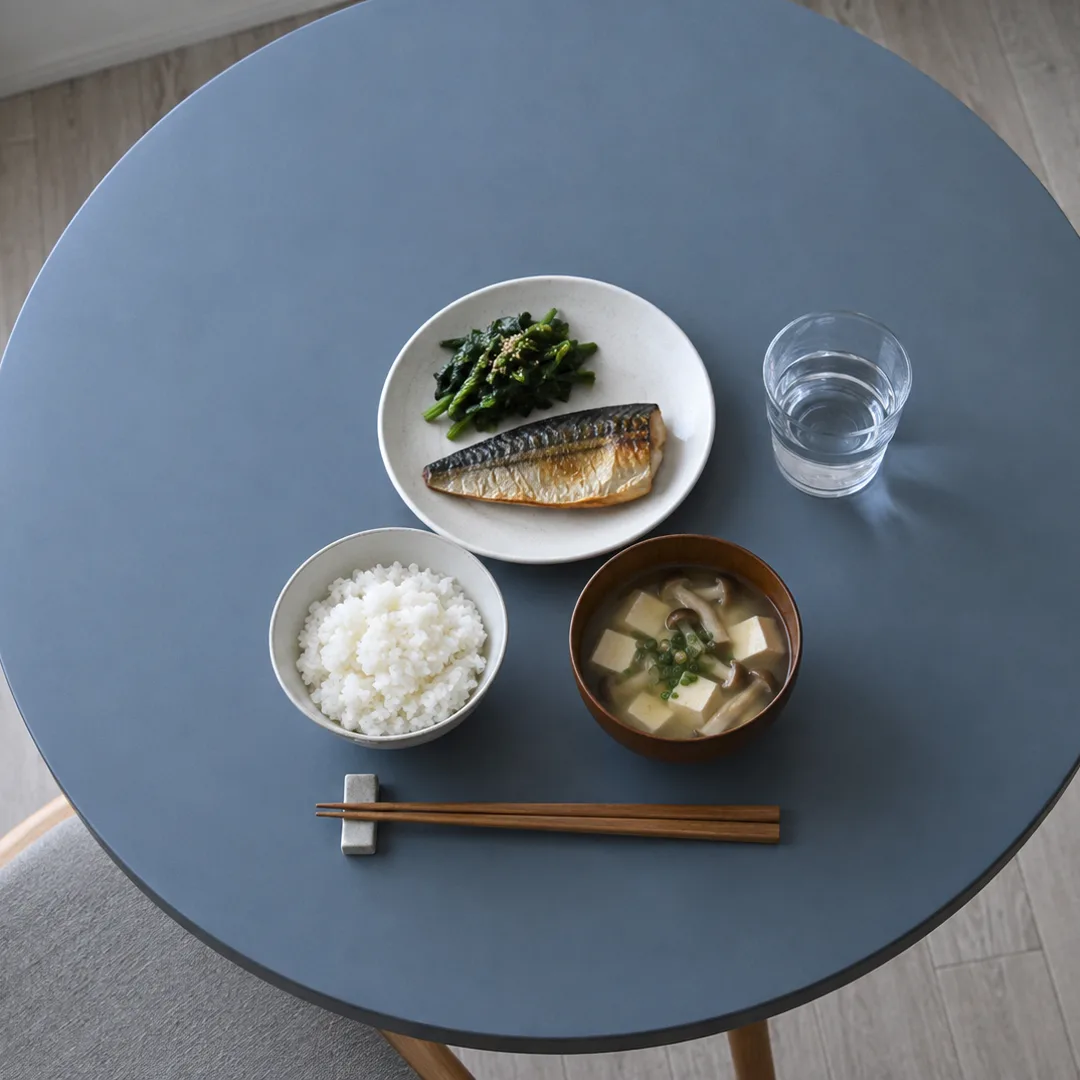 青灰色の丸い食卓に、ご飯、焼き魚、ほうれん草、豆腐ときのこの汁物を白い器で並べた昼食。
