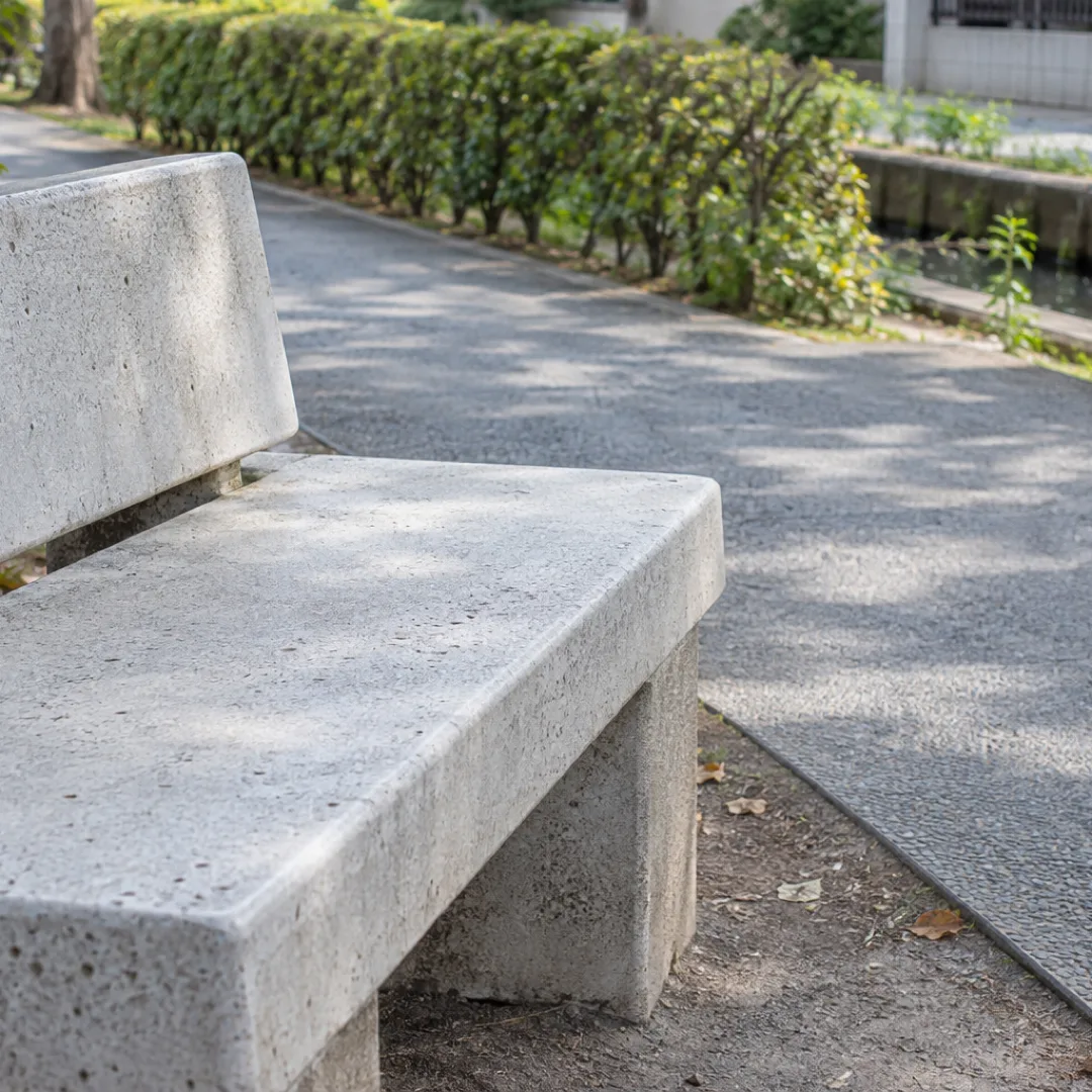 曇り空の柔らかな光の下、日本の小さな公園で平らな歩道の脇に白っぽいコンクリートのベンチがある。