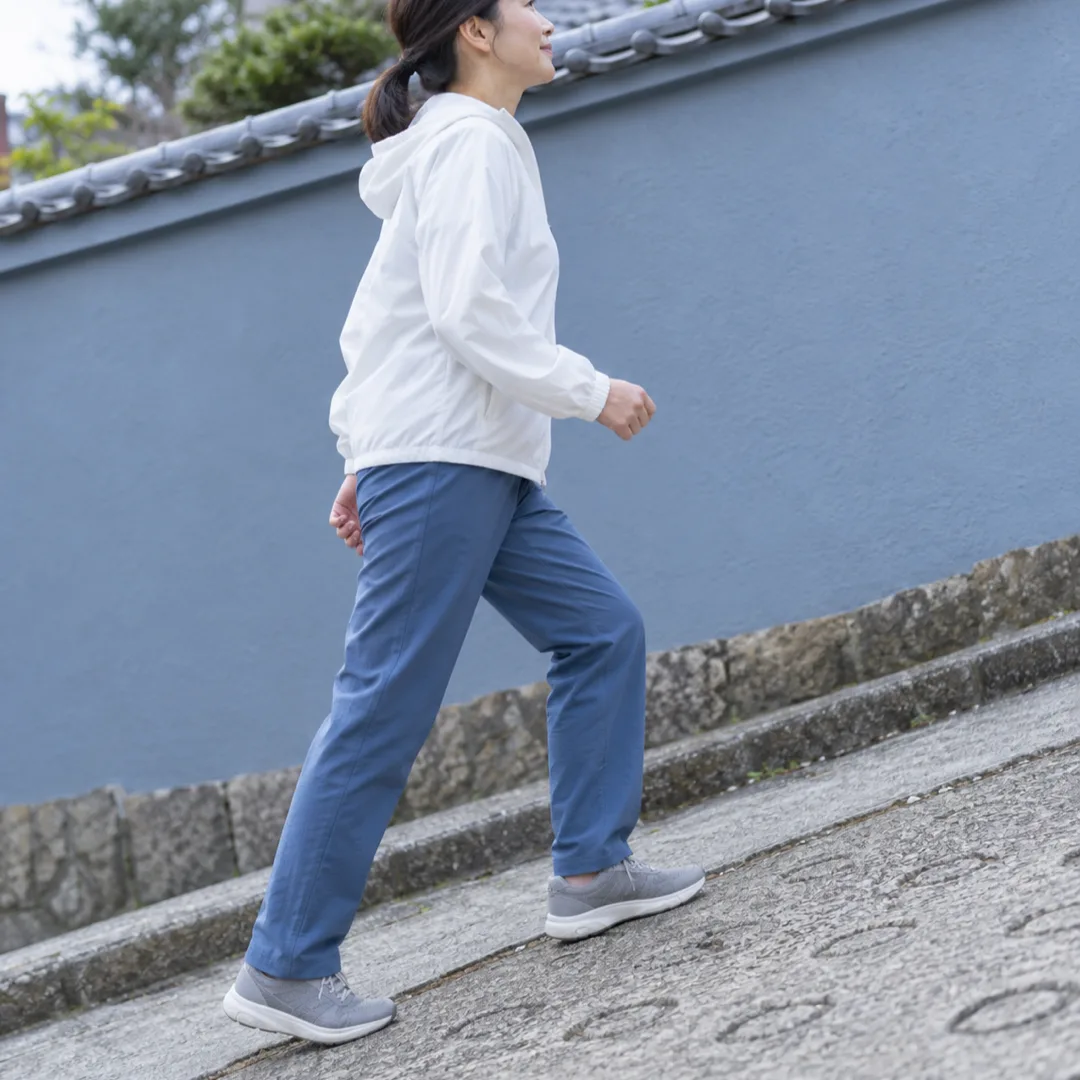 青いズボンと白い上着を着た大人が、日本の住宅街の坂道を小さな歩幅で上る横姿。