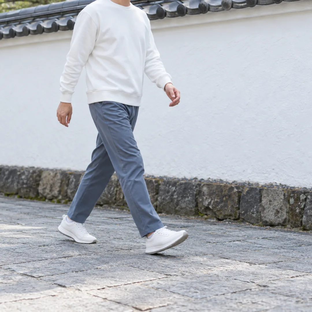 白い塀のある中庭で、白い上着の大人が肘を軽く曲げて歩く、肩から足元までの様子。