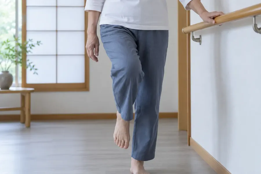 日本の住まいで、白い上着と青灰色のズボンを着た大人が、壁の手すりに片手を添えて裸足の片足を少し浮かせている。