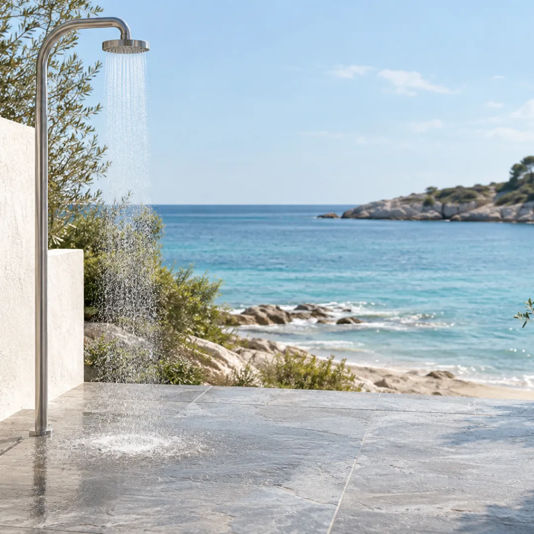 Water falls from an outdoor beach shower onto pale stone, with the aqua sea beyond soft white walls.