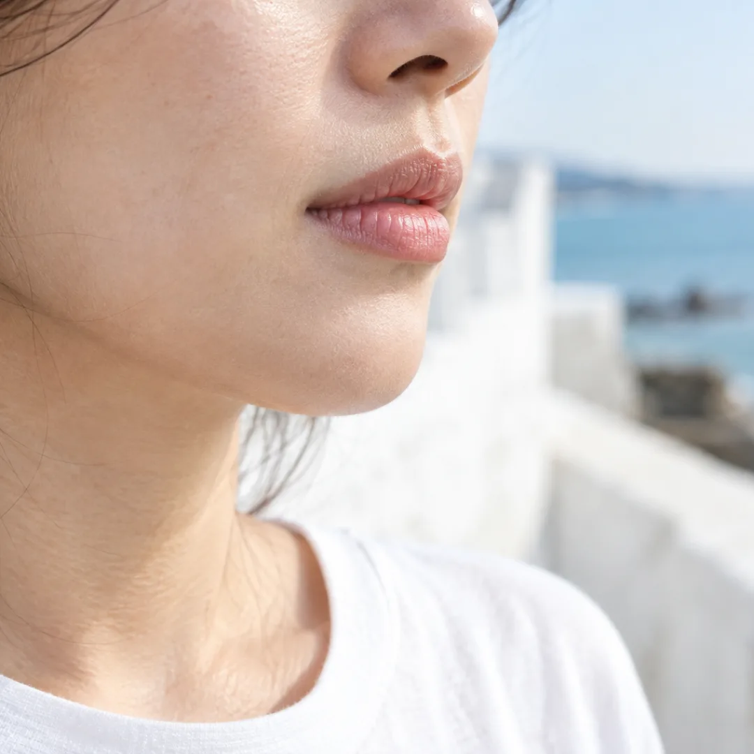 Close-up of an adult’s bare lips and chin in coastal daylight, with a white shirt and pale aqua sea behind.