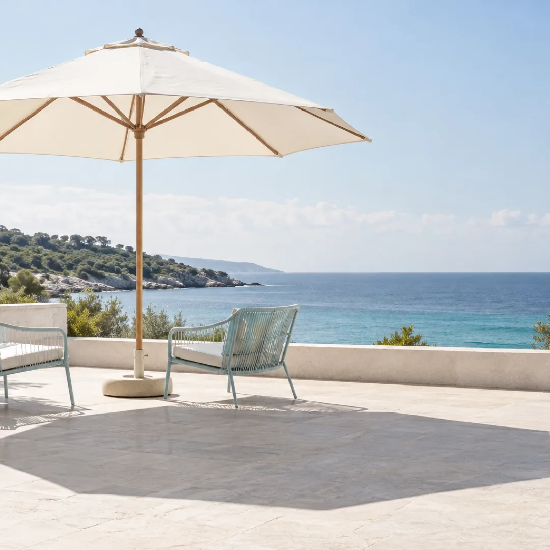 A white parasol shades two light-aqua chairs on an empty terrace overlooking the sea.