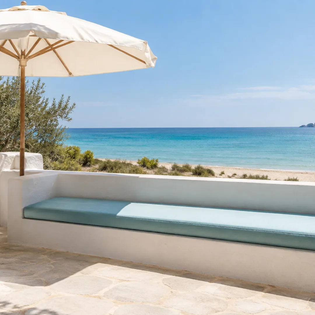 A white seaside terrace with an aqua-cushioned bench partly shaded by a parasol and partly in sunlight.