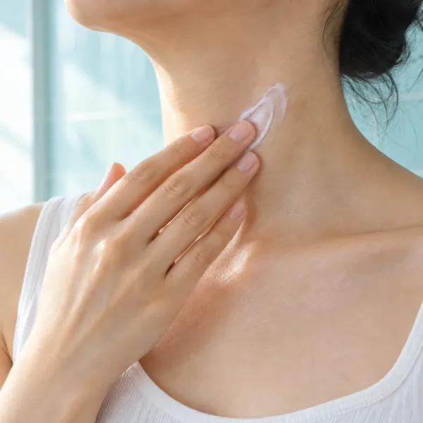 An adult gently spreads moisturizer over the side of their neck against a light aqua bathroom wall.