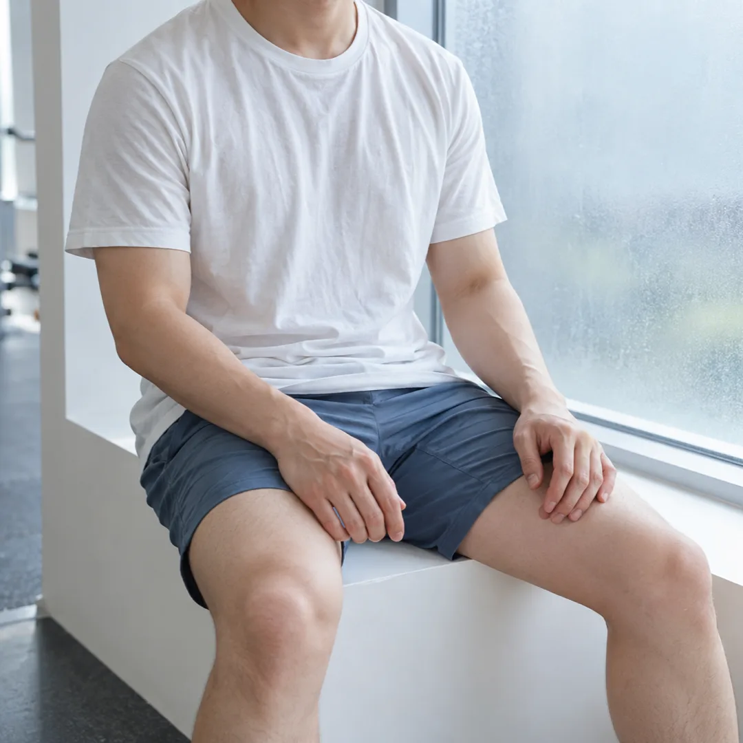 흰 티셔츠와 푸른 운동 반바지를 입은 성인이 체육관 창가에 앉아 두 손을 허벅지에 얹고 쉬고 있다.