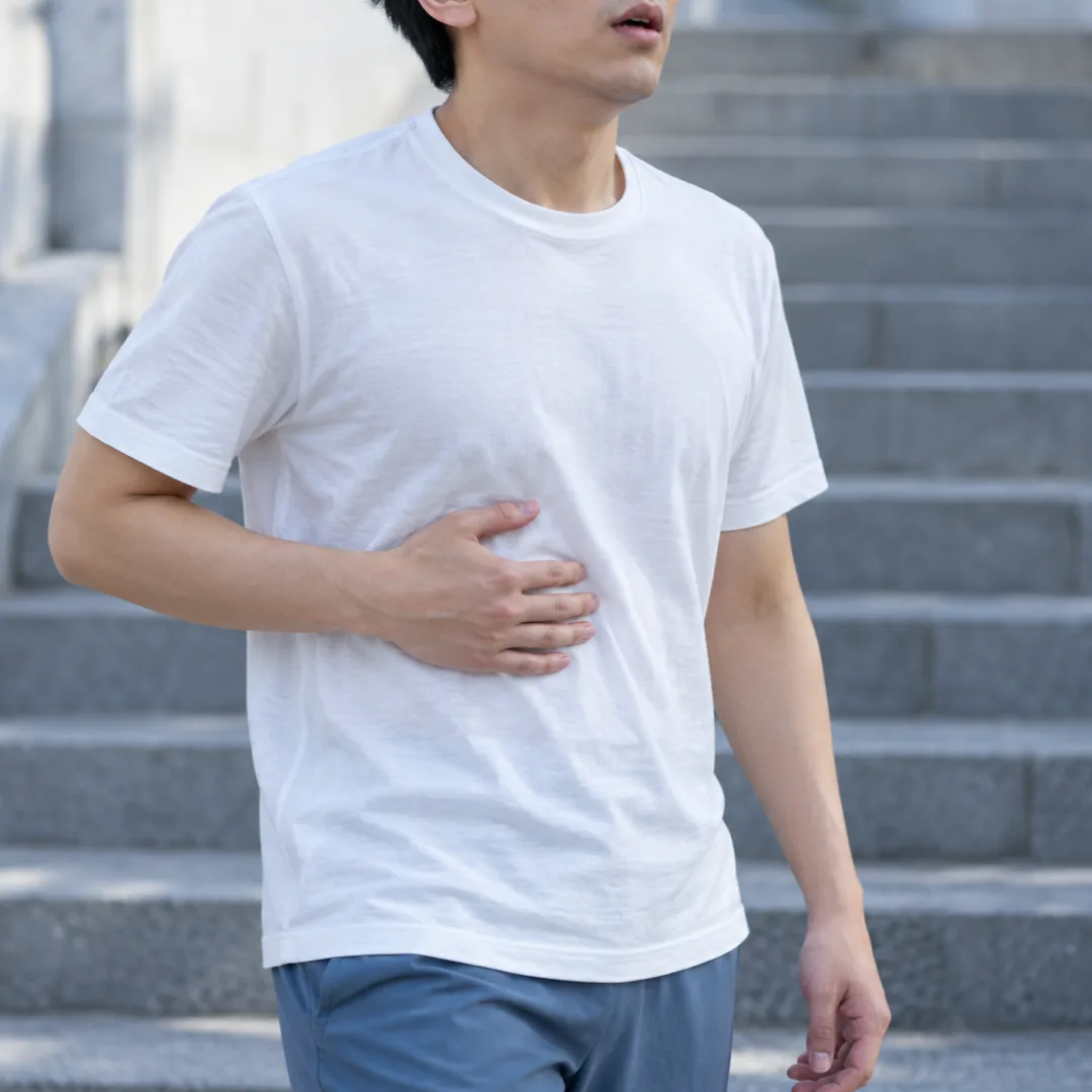 야외 계단 앞에서 흰 운동복을 입은 성인이 한 손을 갈비뼈 아래에 가볍게 대고 서 있는 모습.