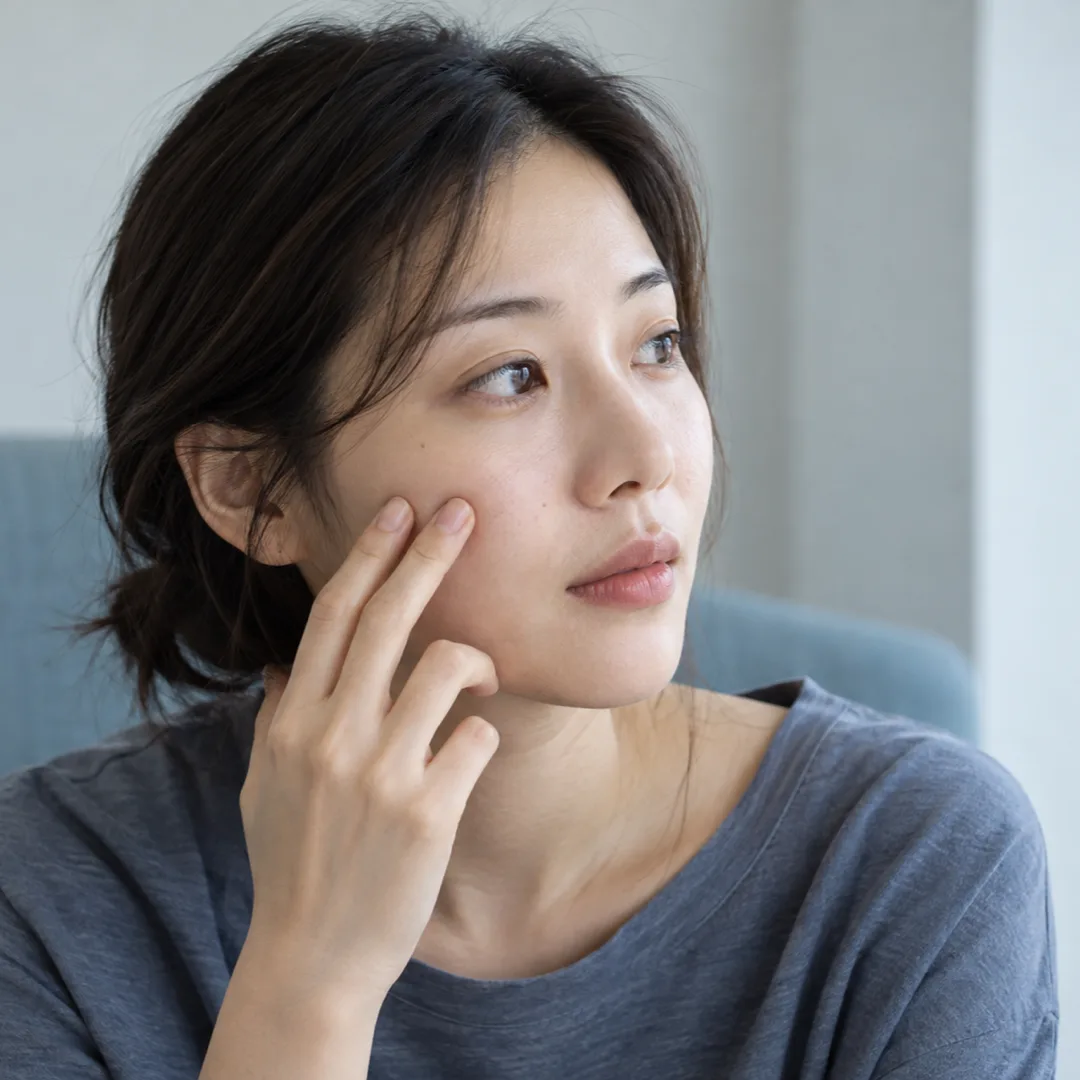A woman in a grey-blue top lightly touches her cheek while seated in a softly lit consultation room.