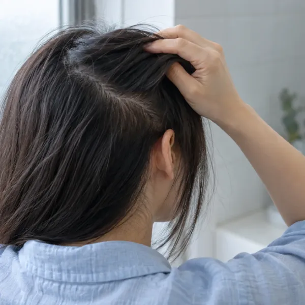 An adult gently lifts a section of dark hair, revealing short strands near the parting beside a frosted window.