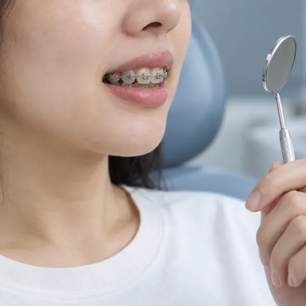 An adult in a dental chair holds a small mirror near their mouth to inspect teeth fitted with metal braces.